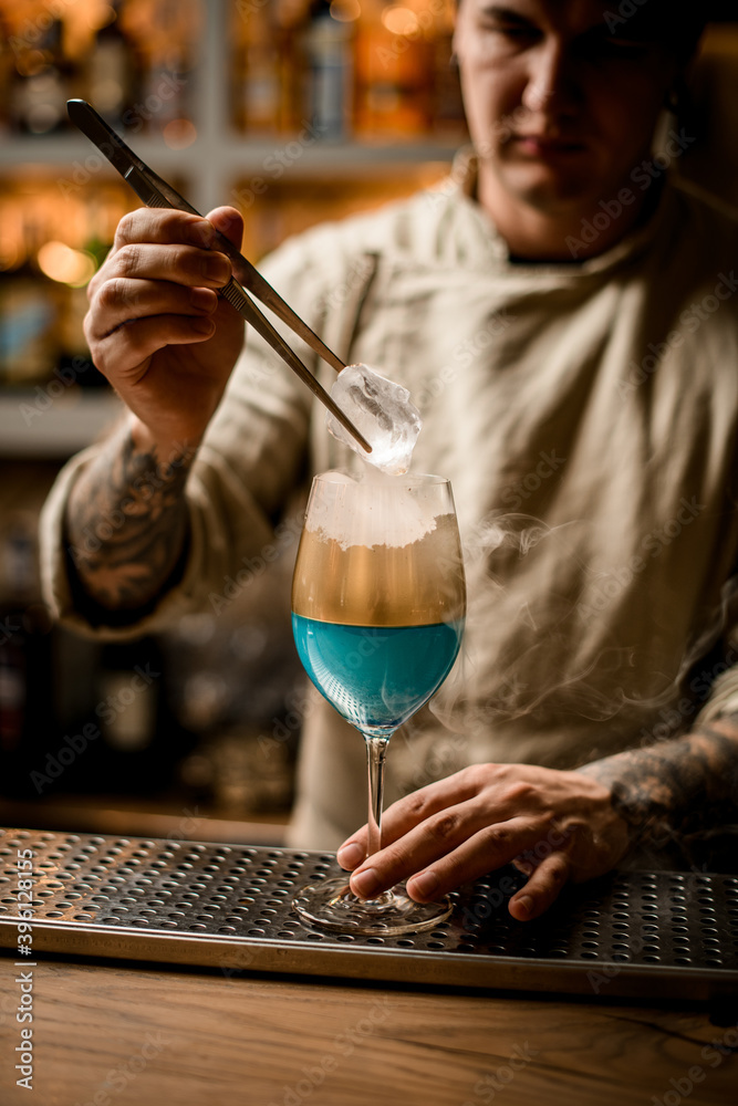 barman holds piece of ice over steaming wine glass decorated with gold