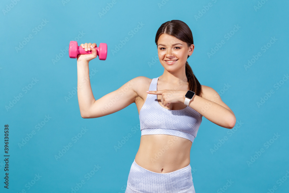 Positive athletic woman in white sportswear pointing finger at pink dumbbell she holding in hand, bragging with biceps, doing workout exercises. Indoor studio shot isolated on blue background Positive athletic woman in white sportswear pointing finger at pink dumbbell she holding in hand, bragging with biceps, doing workout exercises. Indoor studio shot isolated on blue background