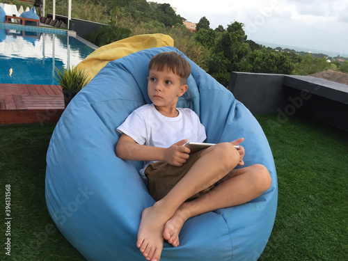 Pensive boy sitting in blue armchair with tablet