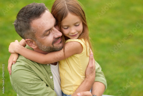 Portrait of happy young father and adorable little daughter hugging each other, standing with eyes closed while spending time in the green park on a summer day © Kostiantyn