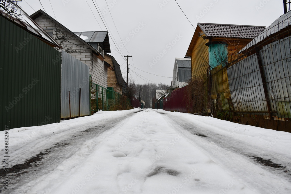 Street in a horticultural Association in late autumn. High metal fences. There is snow on the road. On the horizon is a bare birch forest. Country houses behind high fences. Street in a horticultural Association in late autumn. High metal fences. There is snow on the road. On the horizon is a bare birch forest. Country houses behind high fences.