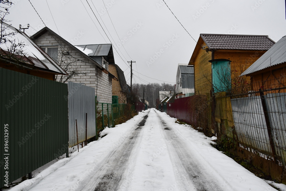 Street in a horticultural Association in late autumn. High metal fences. There is snow on the road. On the horizon is a bare birch forest. Country houses behind high fences. Street in a horticultural Association in late autumn. High metal fences. There is snow on the road. On the horizon is a bare birch forest. Country houses behind high fences.