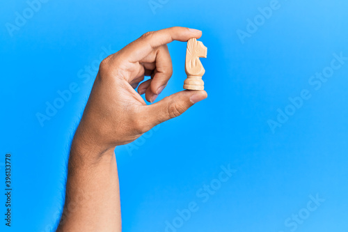 Hand of hispanic man holding horse chess piece over isolated blue background.