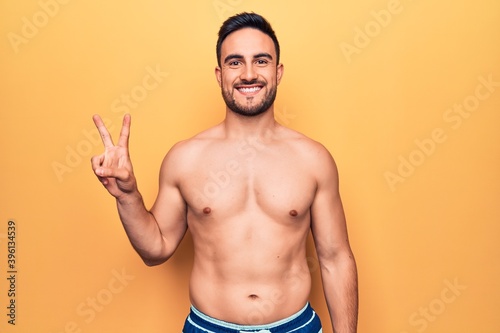 Young handsome man with beard wearing sleeveless t-shirt standing over yellow background smiling with happy face winking at the camera doing victory sign. Number two.