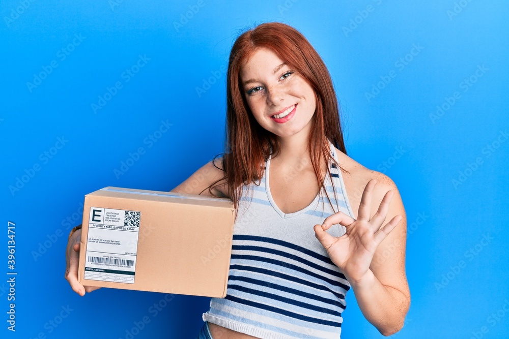 Young red head girl holding delivery package doing ok sign with fingers, smiling friendly gesturing excellent symbol Young red head girl holding delivery package doing ok sign with fingers, smiling friendly gesturing excellent symbol