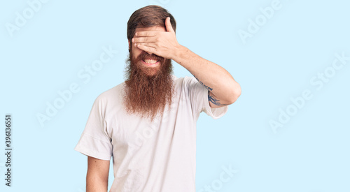 Handsome young red head man with long beard wearing casual white tshirt smiling and laughing with hand on face covering eyes for surprise. blind concept.