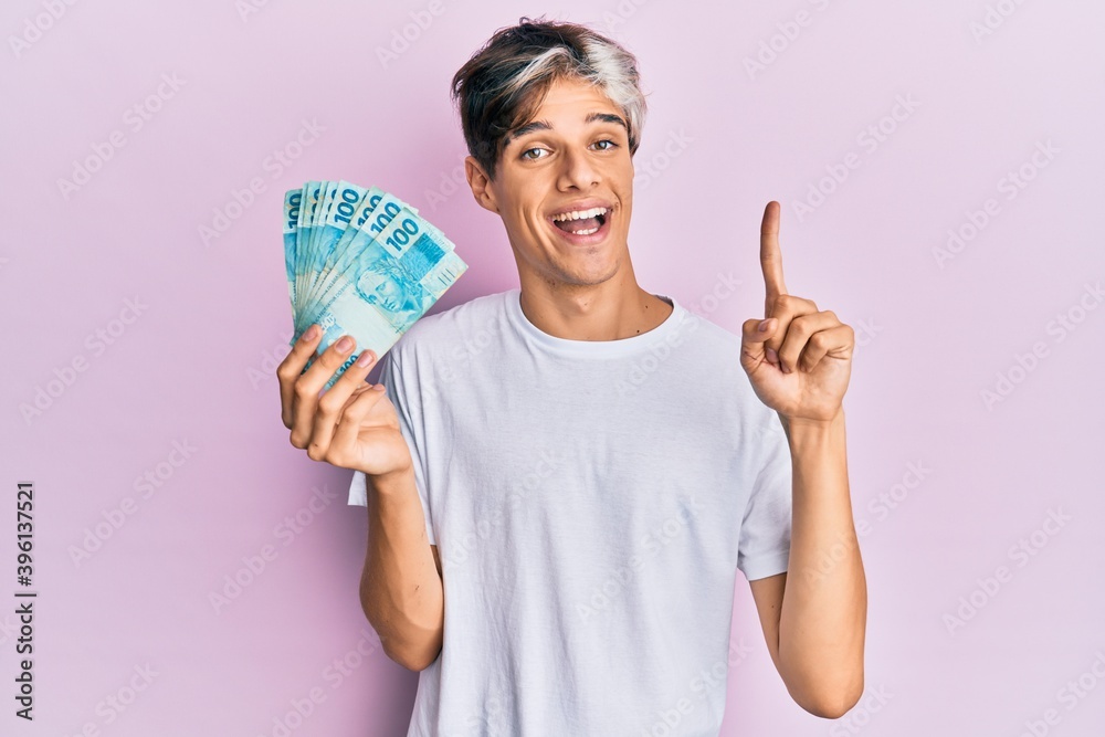Young hispanic man holding 100 brazilian real banknotes smiling with an idea or question pointing finger with happy face, number one Young hispanic man holding 100 brazilian real banknotes smiling with an idea or question pointing finger with happy face, number one