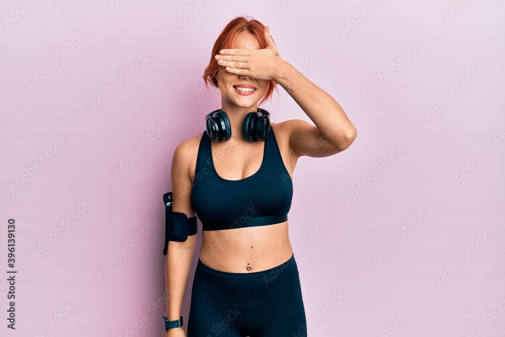 Obraz premium Young beautiful redhead woman wearing gym clothes and using headphones smiling and laughing with hand on face covering eyes for surprise. blind concept.
