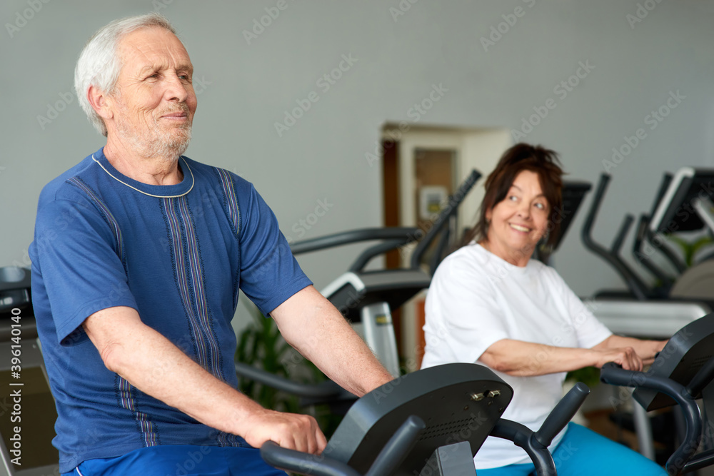 Obraz premium Senior couple training at machine at gym. Elderly cheerful man and woman in gym doing cardio work out, focus on man.