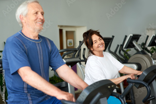 Senior couple spinning on fitness bikes. Happy elderly woman training on machine at gym.