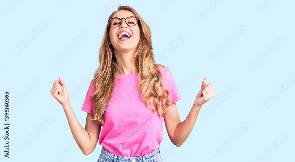 Young beautiful caucasian woman with blond hair wearing casual clothes and glasses celebrating surprised and amazed for success with arms raised and open eyes. winner concept. Young beautiful caucasian woman with blond hair wearing casual clothes and glasses celebrating surprised and amazed for success with arms raised and open eyes. winner concept.