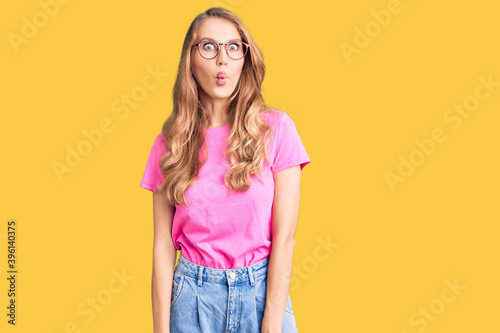 Young beautiful caucasian woman with blond hair wearing casual clothes and glasses making fish face with lips, crazy and comical gesture. funny expression.
