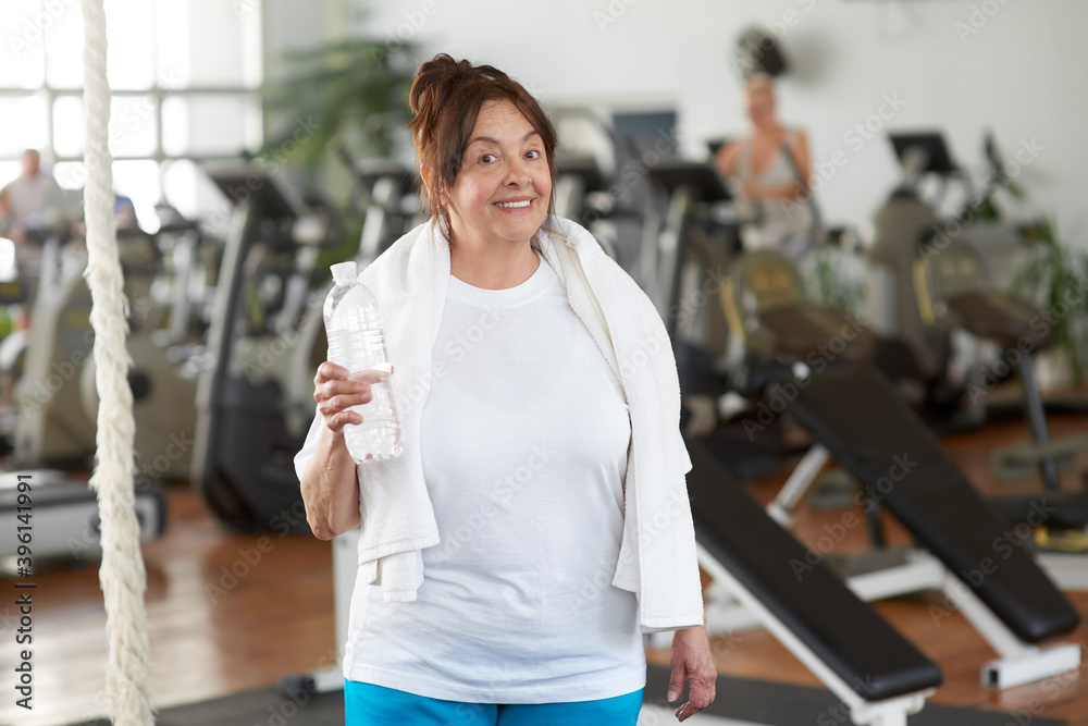 Senior woman holding bottle of water at gym. Happy elderly woman looking at camera at sport club. People, sport and healthy lifestyle.