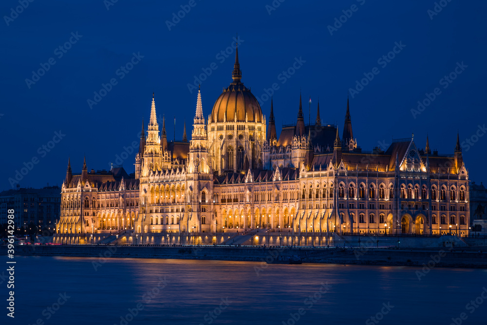 Fototapeta premium A spectacularly illuminated grandiose neo-Gothic building on the river embankment in a European city. Blue hour, clear dark blue sky. No people