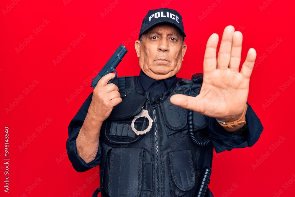 Senior man with grey hair wearing police uniform holding gun with open hand doing stop sign with serious and confident expression, defense gesture Senior man with grey hair wearing police uniform holding gun with open hand doing stop sign with serious and confident expression, defense gesture