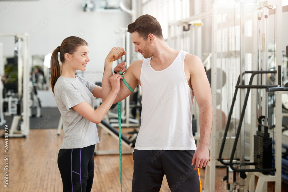 Young sporty man and woman at gym. Pretty smiling young woman measuring the biceps of handsome muscular man. Strength training result. Young sporty man and woman at gym. Pretty smiling young woman measuring the biceps of handsome muscular man. Strength training result.