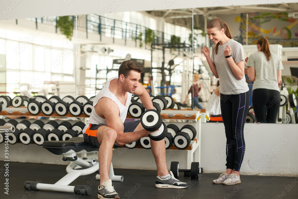 Young beautiful sporty couple at gym. Cheerful young woman looking at male athlete lifting dumbbell at fitness studio. People, sport, relationship and healthy lifestyle concept. Young beautiful sporty couple at gym. Cheerful young woman looking at male athlete lifting dumbbell at fitness studio. People, sport, relationship and healthy lifestyle concept.