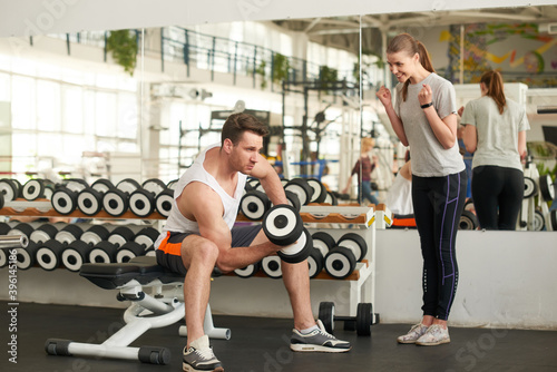 Fototapeta Naklejka Na Ścianę i Meble -  Young beautiful sporty couple at gym. Cheerful young woman looking at male athlete lifting dumbbell at fitness studio. People, sport, relationship and healthy lifestyle concept.