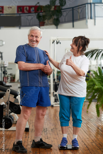 Happy senior couple at gym. Mature woman on weight scale celebrating weightloss progress. People, sport, diet, weight loss and gestures concept.