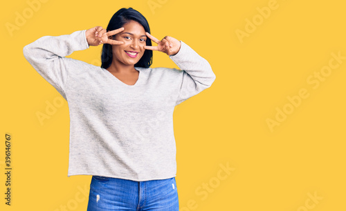 Hispanic woman with long hair wearing casual clothes doing peace symbol with fingers over face, smiling cheerful showing victory