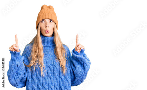 Beautiful caucasian woman with blonde hair wearing wool sweater and winter hat amazed and surprised looking up and pointing with fingers and raised arms. photo