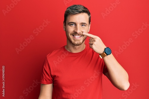 Handsome caucasian man wearing casual red tshirt pointing with hand finger to face and nose, smiling cheerful. beauty concept