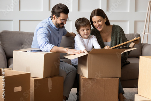 Happy young Caucasian family with small son unpack cardboard boxes with personal belongings moving to new home. Excited parents with little child settle unbox relocating to own house. Rental concept.