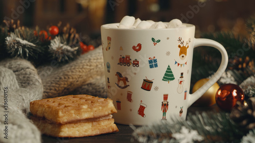 Hot cocoa with marshmallows, cinnamon, Viennese waffle in white mug, surrounded by winter Christmas tree branches with berries and cones on a wooden table. The concept of a cozy holiday and New Year