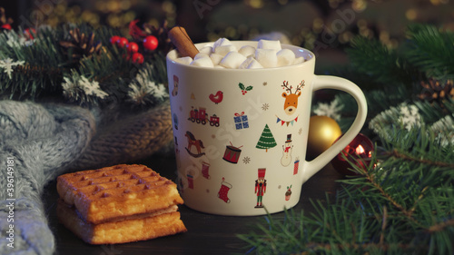 Hot cocoa with marshmallows, cinnamon, Viennese waffle in white mug, surrounded by winter Christmas tree branches with berries and cones on a wooden table. The concept of a cozy holiday and New Year