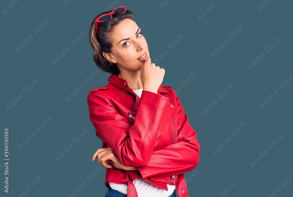 Young beautiful woman wearing red leather jacket looking confident at the camera with smile with crossed arms and hand raised on chin. thinking positive.