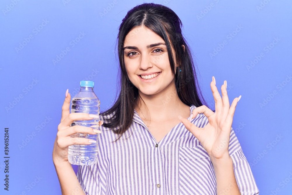 Young beautiful girl holding bottle of water doing ok sign with fingers, smiling friendly gesturing excellent symbol Young beautiful girl holding bottle of water doing ok sign with fingers, smiling friendly gesturing excellent symbol