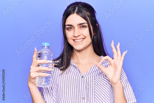 Young beautiful girl holding bottle of water doing ok sign with fingers, smiling friendly gesturing excellent symbol