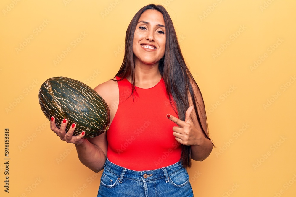 Young beautiful brunette woman holding melon smiling happy pointing with hand and finger Young beautiful brunette woman holding melon smiling happy pointing with hand and finger