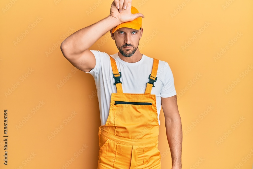 Young handsome man wearing handyman uniform over yellow background making fun of people with fingers on forehead doing loser gesture mocking and insulting.