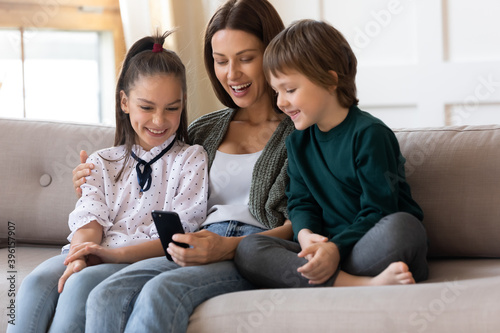 Smiling young Caucasian mother and two small children sit res on sofa in living room look at cellphone screen watch funny video together. Happy mom with kids relax at home using modern smartphone.