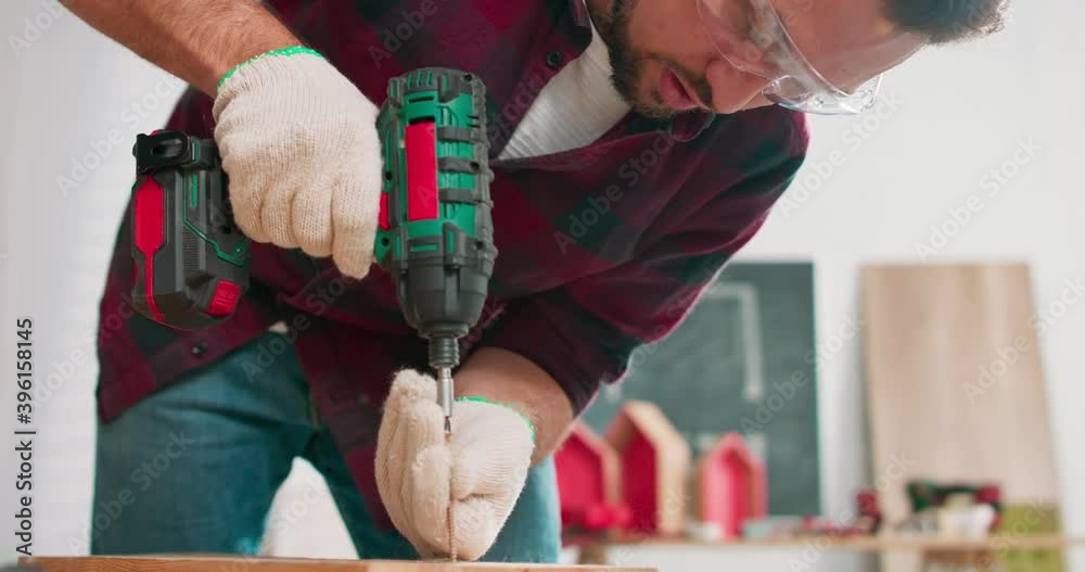 Concentrated young bearded carpenter wearing jeans and a plaid flannel shirt drilling a hole in a piece of wood with a portable electric drill in his carpentry workshop.