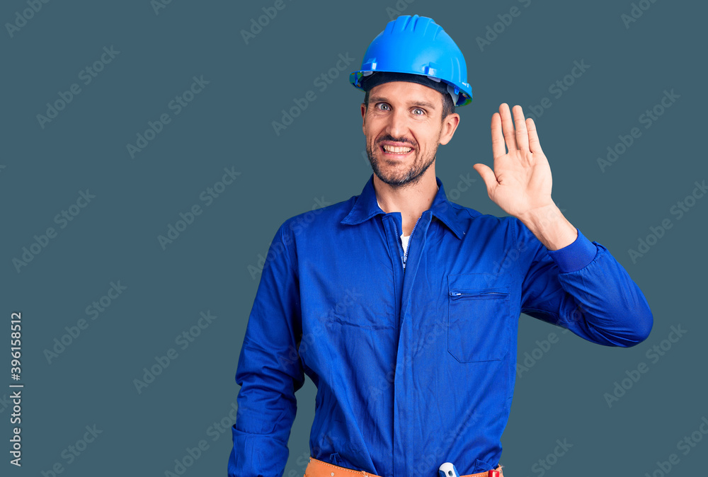 Young handsome man wearing worker uniform and hardhat waiving saying hello happy and smiling, friendly welcome gesture Young handsome man wearing worker uniform and hardhat waiving saying hello happy and smiling, friendly welcome gesture