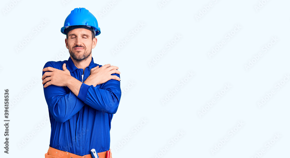 Young handsome man wearing worker uniform and hardhat hugging oneself happy and positive, smiling confident. self love and self care