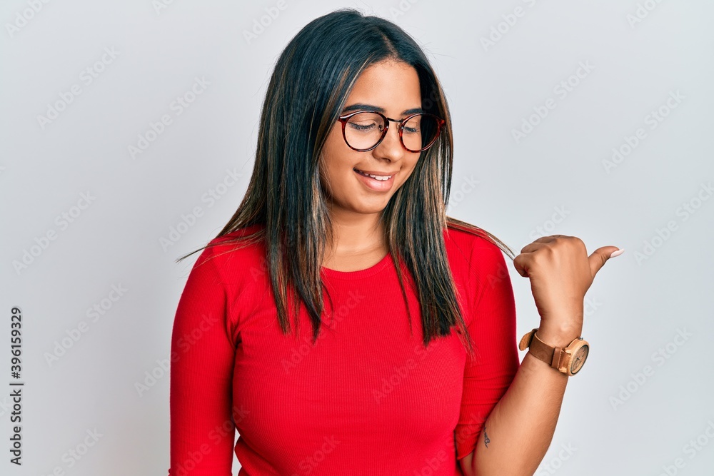 Young latin girl wearing casual clothes and glasses pointing thumb up to the side smiling happy with open mouth Young latin girl wearing casual clothes and glasses pointing thumb up to the side smiling happy with open mouth