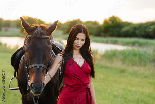 A young pretty girl in a red dress poses on a ranch with a thoroughbred stallion at sunset. Love and care for animals