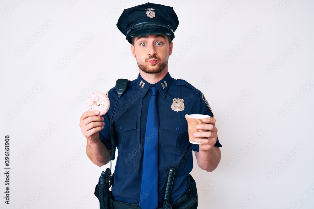 Young caucasian man wearing police uniform holding take away coffee and donut puffing cheeks with funny face. mouth inflated with air, catching air.