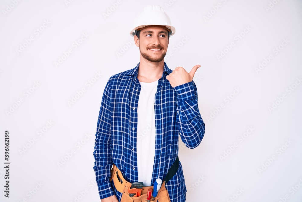Young handsome man weaing handyman uniform smiling with happy face looking and pointing to the side with thumb up. Young handsome man weaing handyman uniform smiling with happy face looking and pointing to the side with thumb up.