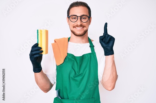 Young handsome man wearing apron holding scourer surprised with an idea or question pointing finger with happy face, number one