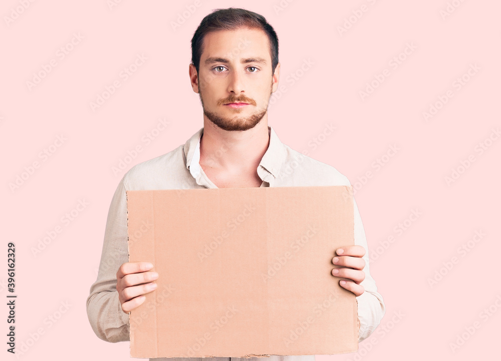 Young handsome caucasian man holding empty cardboard banner thinking attitude and sober expression looking self confident Young handsome caucasian man holding empty cardboard banner thinking attitude and sober expression looking self confident