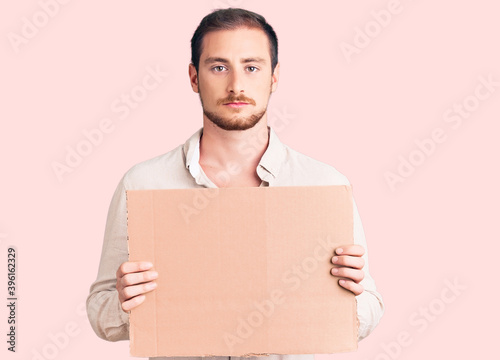 Young handsome caucasian man holding empty cardboard banner thinking attitude and sober expression looking self confident