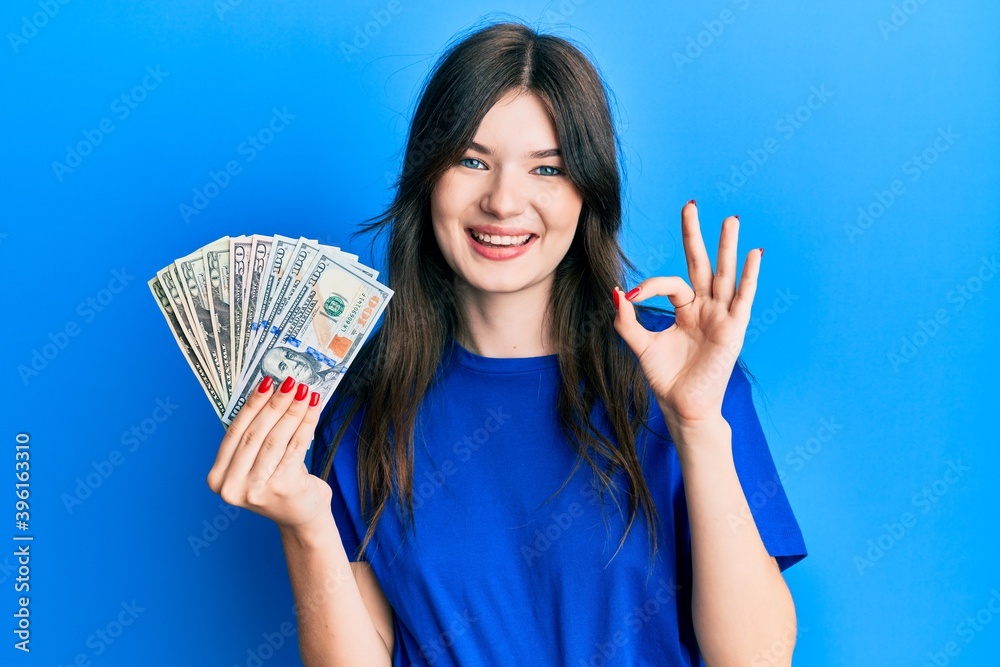 Young beautiful caucasian girl holding dollars doing ok sign with fingers, smiling friendly gesturing excellent symbol Young beautiful caucasian girl holding dollars doing ok sign with fingers, smiling friendly gesturing excellent symbol