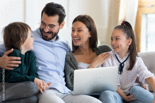 Overjoyed young Caucasian family renters with children sit rest on couch at home use modern laptop gadget together. Smiling parents with small kids relax on sofa in living room browse on computer.