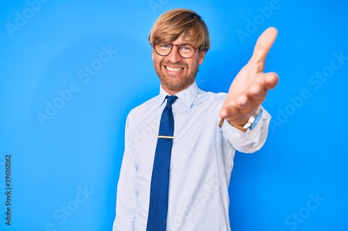 Young blond man wearing business clothes and glasses smiling friendly offering handshake as greeting and welcoming. successful business.