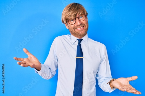 Young blond man wearing business clothes and glasses clueless and confused expression with arms and hands raised. doubt concept.