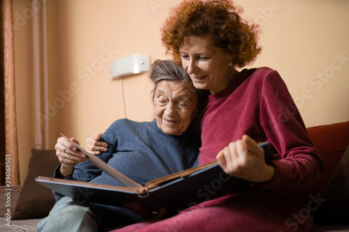 Senior woman and her adult daughter looking at photo album together on couch in living room, talking joyful discussing memories. Weekend with parents, family day, thanksgiving, mom's holiday